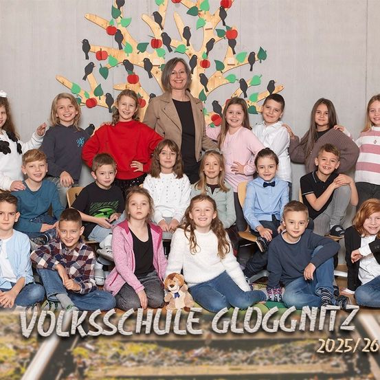 Ein Gruppenfoto von Schülern und einer Lehrerin an der Volksschule Gloggnitzer. Sie sitzen in Reihen und lächeln. Die Lehrerin steht in der Mitte und hinter ihnen ist ein dekorierter Apfelbaum. Das Bild hat einen Wasserzeichen mit dem Text '2025/26'.