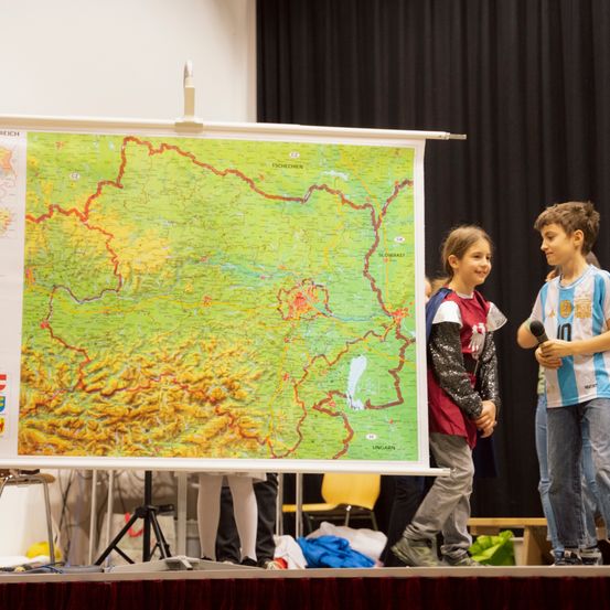 Zwei Kinder stehen auf der Bühne, eines hält ein Mikrofon und das andere steht neben einer großen Karte. Hinter ihnen befindet sich ein schwarzer Hintergrund und eine Person, die auf einem Stuhl sitzt.