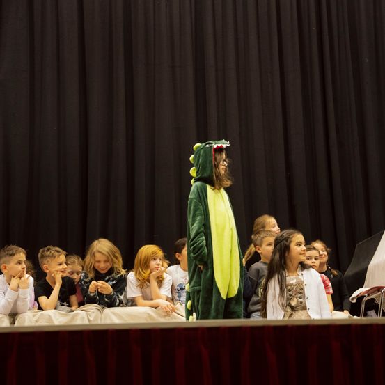 Eine Gruppe von Kindern beobachtet eine Person in einer grünen Dinosaurier-Kostümierung auf der Bühne. Einige Kinder sitzen mit den Händen vor dem Mund.