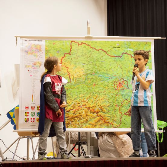 Zwei Jungen stehen auf der Bühne, einer mit einem Mikrofon. Einer ist in mittelalterlicher Kleidung. Sie stehen vor einer großen Karte einer Region.