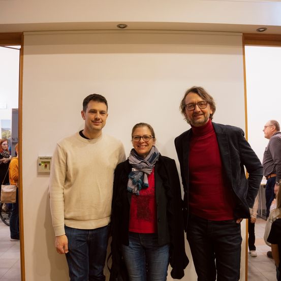 Drei Personen posieren für ein Foto in einem Innenraum, mit einer weißen Wand und einer offenen Tür im Hintergrund. Der Mann links trägt einen beigen Pullover, die Frau in der Mitte trägt einen Schal und eine Brille, und der Mann rechts trägt ein rotes Rollkragenshirt und eine Brille.