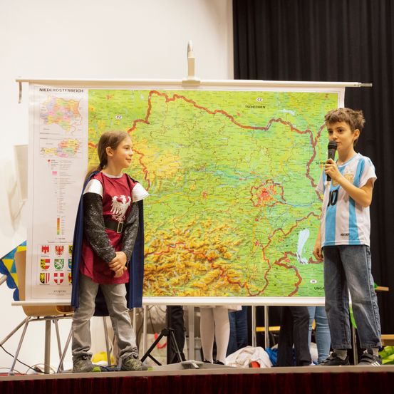 Zwei Kinder stehen auf der Bühne, eines hält ein Mikrofon, und sie stehen vor einer großen Karte von Niederösterreich.