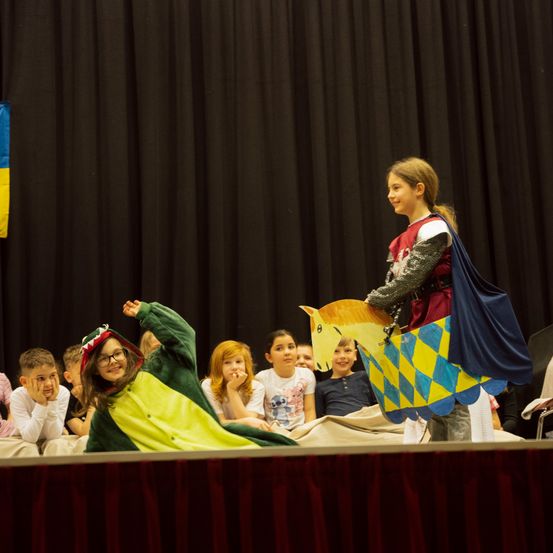 Kinder in Kostümen spielen auf der Bühne. Ein Kind ist als Ritter verkleidet und hält ein Spielzeugpferd, während ein anderes Kind als Drache auf der Bühne liegt.
