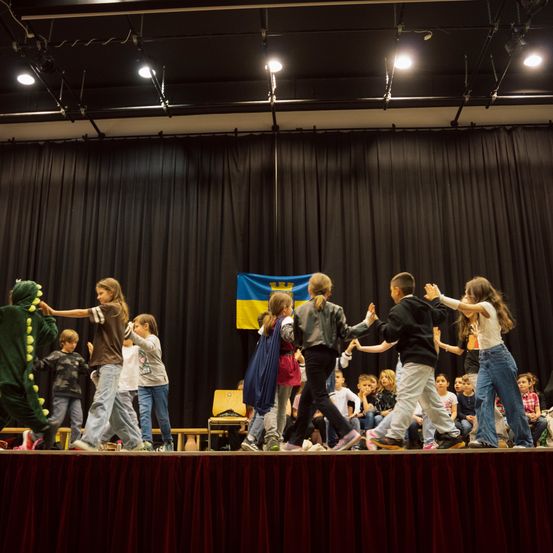 Eine Gruppe von Kindern steht auf der Bühne und führt ein Theaterstück mit Kostümen auf, eines als Dinosaurier verkleidet. Ein Flaggenhintergrund ist zu sehen.