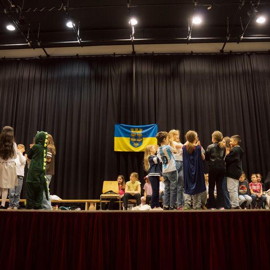 Eine Gruppe von Kindern steht auf der Bühne mit einer Flagge dahinter. Einige sind in Kostümen gekleidet, während andere von unten zuschauen.
