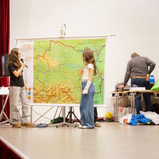 Zwei Frauen stehen auf einer Bühne vor einer großen Karte, eine spricht in ein Mikrofon. Ein Mann im Hintergrund beugt sich über einen Tisch mit verschiedenen Gegenständen.