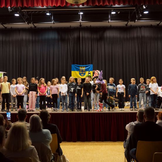 Eine Gruppe von Kindern steht auf der Bühne, einige halten Besen. Eine Flagge hängt hinter ihnen. Ein Publikum sitzt vor der Bühne.