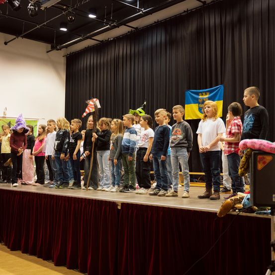 Eine Gruppe von Kindern steht auf der Bühne eines Theaters, trägt Kostüme und Requisiten. Ein Flagge ist im Hintergrund sichtbar.