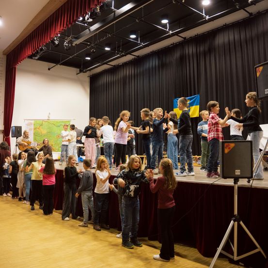 Eine Gruppe von Kindern und Erwachsenen tanzt auf einer Bühne in einem Theater. Eine Person spielt Gitarre. Eine Flagge und eine Karte sind auf der Bühne.