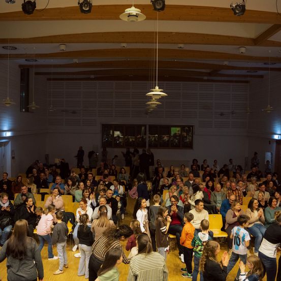 Eine große Menschenmenge versammelt sich in einem Saal, klatscht und lächelt, einige stehen und andere sitzen in Reihen von Stühlen. Der Saal hat eine Holzdecke und weiße Wände, mit Lampen, die daran hängen.