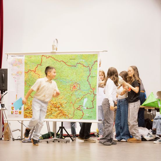 Eine Gruppe von Kindern steht um eine große Karte auf einem Ständer herum. Ein Junge läuft an ihnen vorbei. Hinter der Karte stehen mehrere Personen.