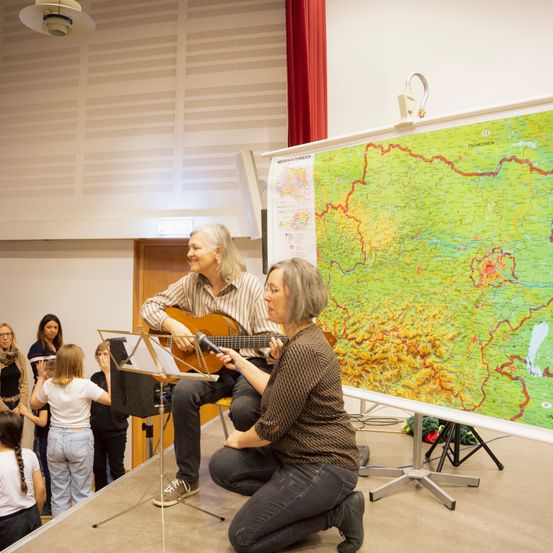 Eine Frau interviewt einen Mann, der Gitarre auf der Bühne spielt, in einem Klassenzimmer mit einer großen Karte im Hintergrund. Kinder schauen zu.