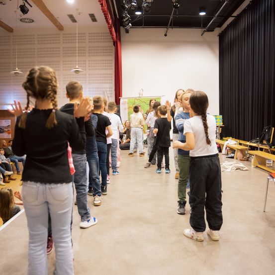 Eine Gruppe von Kindern und Erwachsenen steht in einem Raum, einige klatschen. Eine Karte ist an der Wand. Eine Person in Weiß tanzt vor anderen, während andere zusehen.
