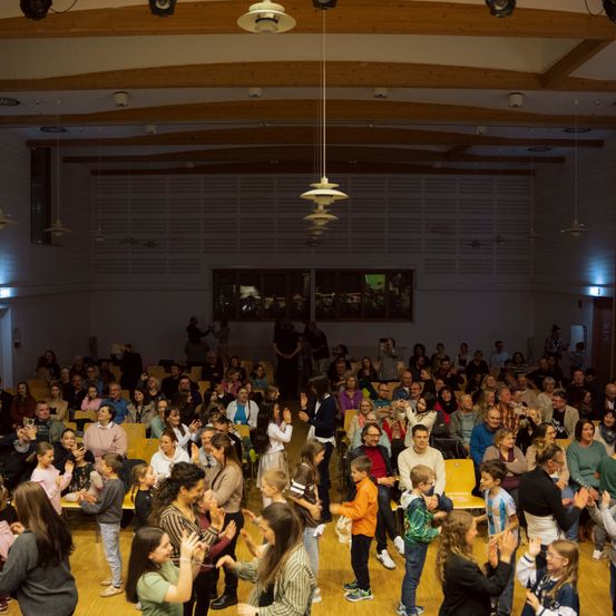 Eine große Gruppe von Menschen unterschiedlichen Alters in einem Saal, die klatschen und lächeln. Der Raum ist gut beleuchtet, mit Holzbalken an der Decke und großen Fenstern. Einige Leute stehen, während andere auf Stühlen sitzen.