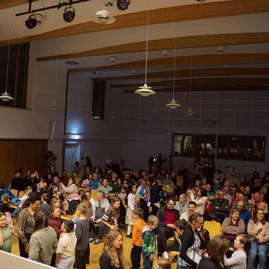 Eine große Versammlung von Menschen in einem Saal, viele stehend und einige sitzend, mit Menschen, die Fotos machen und andere zuschauen. Der Raum hat Holzbalken, hängende Lampen und Glasfenster.
