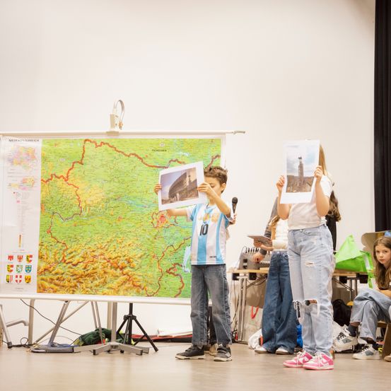 Zwei Kinder stehen auf einer Bühne und halten Bilder. Eines hält ein Mikrofon und eine große Karte hängt hinter ihnen. Andere Leute stehen hinter ihnen, einer hält ein Buch.