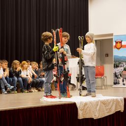 Drei Kinder stehen auf der Bühne und halten Skier. Hinter ihnen sitzen Kinder auf einer Bank und schauen zu. Ein Kind trägt Handschuhe und hält ein Paar Skier. Ein schwarzer Vorhang ist im Hintergrund.