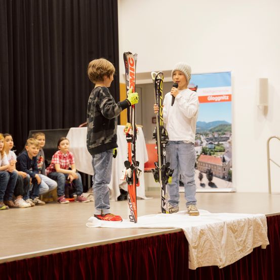 Zwei Kinder auf der Bühne mit Skiern, eines mit Handschuhen, das andere mit einem Mikrofon, vor einem Publikum.