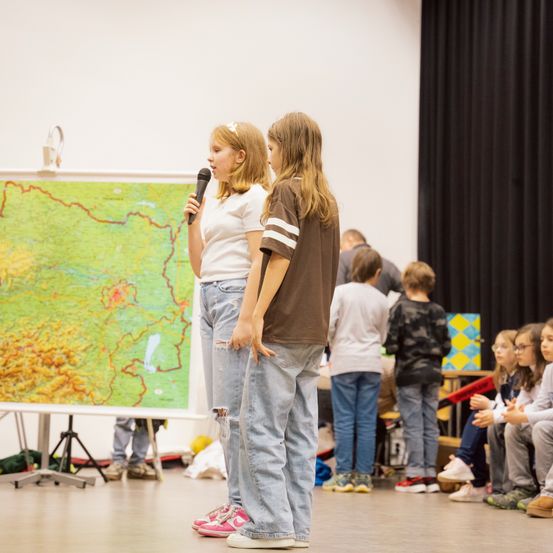 Zwei Mädchen stehen vor einer Karte, eine hält ein Mikrofon. Dahinter sitzen und stehen Kinder in einem Klassenzimmer.