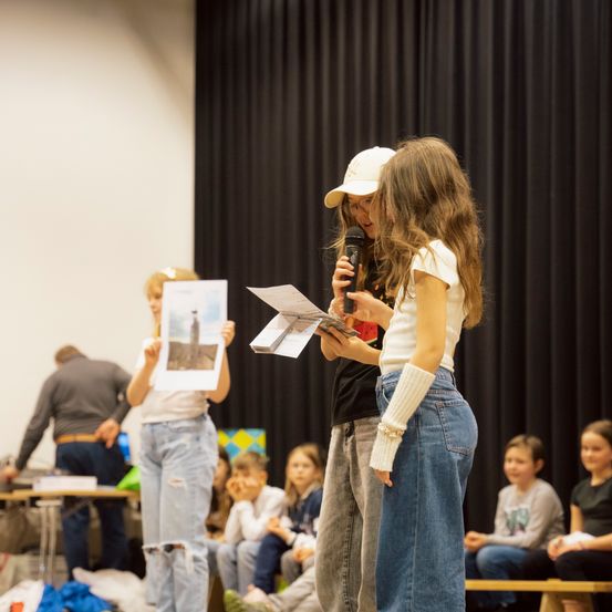Zwei Mädchen stehen auf der Bühne und halten ein Mikrofon und ein Buch. Sie scheinen zu präsentieren oder laut zu lesen. Im Hintergrund sitzt eine Gruppe von Kindern auf einer Bank und schaut aufmerksam zu. Ein Mann steht in der Nähe eines Tisches mit Büchern.