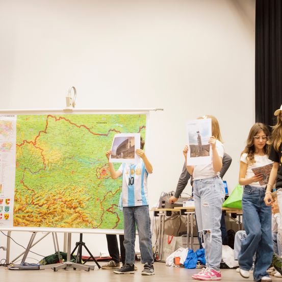 Eine Gruppe von Kindern steht drinnen, hält Fotos in der Hand und sieht auf eine große Karte auf einem Ständer. Sie tragen Freizeitkleidung und haben Bücher in der Hand.
