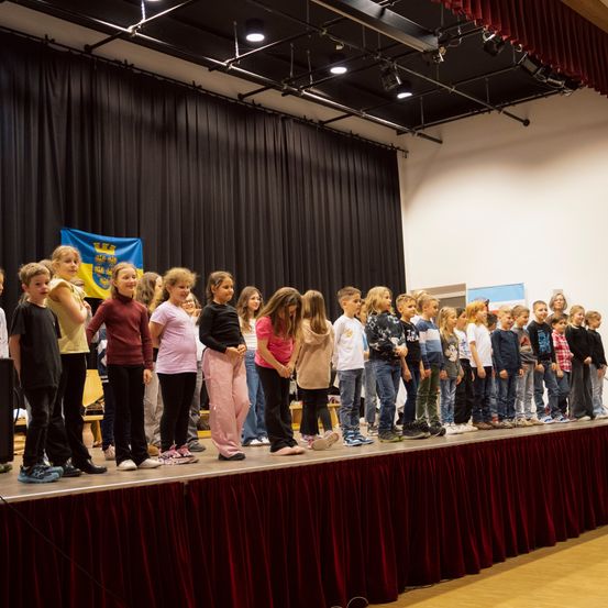 Eine Gruppe von Kindern steht auf der Bühne, wahrscheinlich bei einer Aufführung. Die Bühne ist mit einem roten Vorhang bedeckt, und eine Fahne ist im Hintergrund zu sehen. Der Raum hat einen schwarzen Vorhang und eine Deckenbeleuchtung.