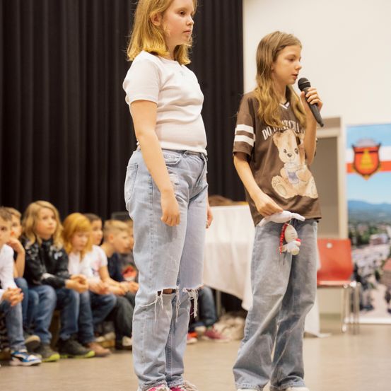 Zwei junge Mädchen stehen auf der Bühne. Eine hält ein Mikrofon, die andere einen Kuscheltier. Kinder sitzen hinter ihnen und schauen zu.