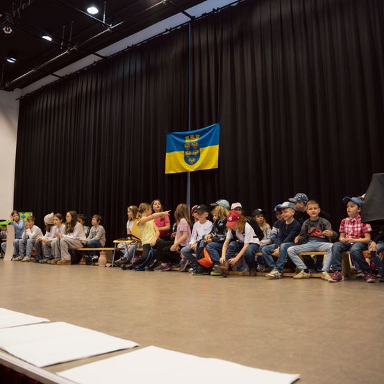 Kinder sitzen auf Bänken auf einer Bühne und schauen auf eine ukrainische Flagge im Hintergrund. Sie tragen verschiedene Kleidungsstücke und Schuhe. Die Bühne ist mit Deckenleuchten beleuchtet.