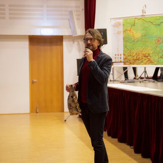 Ein Mann spricht in ein Mikrofon auf einer Bühne, hält eine Tasche und hat eine Karte hinter sich und eine Tür zur Seite.