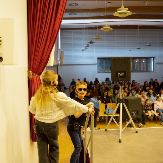 Zwei Kinder, eines mit Kopfhörern, stehen auf der Bühne vor einem Publikum. Die Bühne ist mit einem Mikrofon an einem Stativ ausgestattet. Der Raum hat einen roten Vorhang und Holzdeckenlampen.