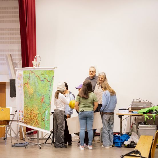 Eine Gruppe von Menschen, darunter zwei Frauen, stehen um eine große Karte in einem Raum. Ein Mann mit Brille steht hinter ihnen. Ein Tisch mit einer Tasche und anderen Gegenständen ist in der Nähe.