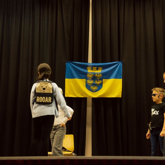 Zwei Personen stehen auf einer Bühne mit einer gelb-blauen Flagge. Ein Mädchen hat einen Rucksack mit dem Wort 'ROOAR' und ein Junge trägt eine Sonnenbrille. Ein Stuhl steht hinter ihnen.
