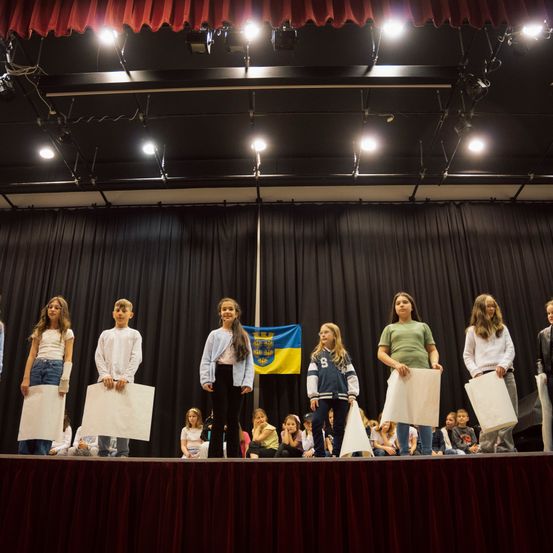 Kinder stehen auf der Bühne und halten Papiere in der Hand. Sie stehen dem Publikum gegenüber. Hinter ihnen befindet sich ein schwarzer Vorhang und die Bühne hat einen roten Vorhang. Im Hintergrund ist eine ukrainische Flagge zu sehen.