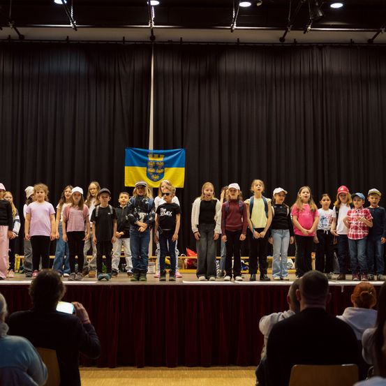 Eine Gruppe von Kindern steht mit einem Publikum auf der Bühne und hält eine blau-gelbe Flagge im Hintergrund.