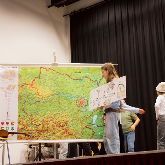 Ein junges Mädchen, das ein Schild hält, steht auf der Bühne und präsentiert vor einer großen Karte. Das Publikum im Hintergrund ist verschwommen.