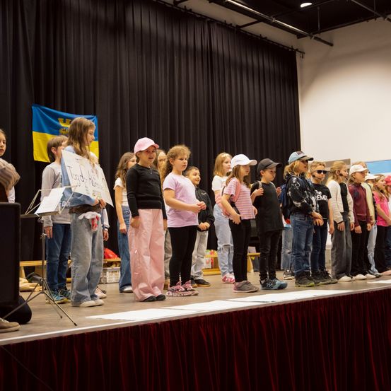 Eine Gruppe von Kindern steht mit einem roten Vorhang auf der Bühne, einige tragen Turnschuhe und Mützen und halten ein Schild. Dahinter weht eine Flagge und ein schwarzer Vorhang.