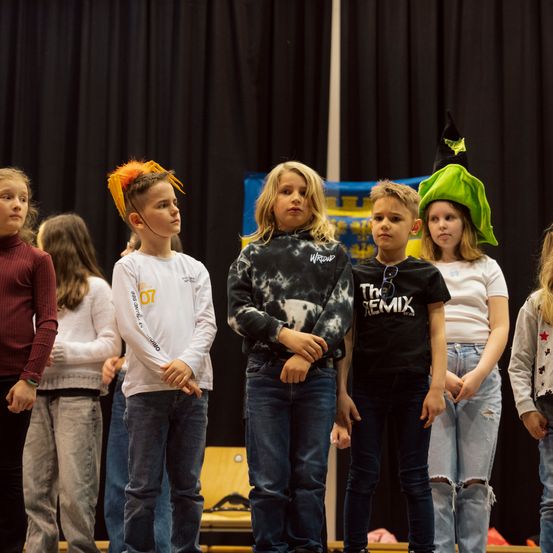 Eine Gruppe von Kindern mit verschiedenen Hüten steht in einer Reihe auf der Bühne. Ein Junge mit orangefarbenen Federn, einem Tie-Dye-Hoodie und einem Mädchen mit einem grünen Hexenhut sind darunter. Sie scheinen auf etwas zu warten.