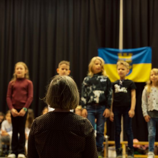 Eine Gruppe von Kindern steht auf einer Bühne mit einer Flagge dahinter, wahrscheinlich bei einer Schulaufführung. Ein Kind mit grauem Haar steht mit dem Rücken zur Bühne und beobachtet die anderen.