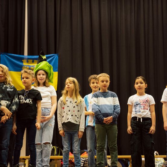 Eine Gruppe von Kindern steht auf einer Bühne vor einem schwarzen Vorhang. Ein Mädchen trägt einen grünen Hexenhut. Sie nehmen wahrscheinlich an einer Schulaufführung teil.