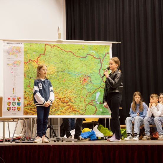 Ein Mädchen mit Mikrofon steht auf der Bühne mit einer Karte im Hintergrund. Ein weiteres Mädchen steht links von ihr, und drei Kinder sitzen auf einer Bank vor ihnen.