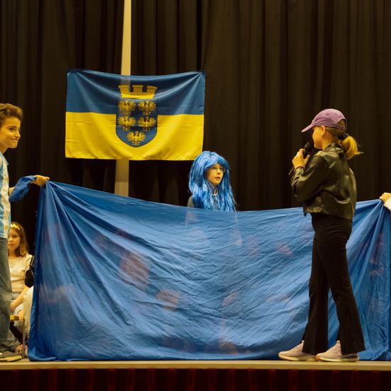 Auf der Bühne führen eine Gruppe von Kindern eine Aufführung mit einer blauen Flagge durch. Ein Kind trägt eine blaue Perücke. Eine Frau mit einer lila Mütze spricht in ein Mikrofon. Dahinter ist eine Flagge mit einem goldenen Adler.
