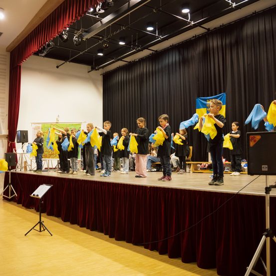 Eine Gruppe Kinder steht mit gelben Flaggen auf der Bühne, trägt schwarze T-Shirts und scheint aufzutreten. Eine Flagge der Ukraine hängt an der Wand hinter ihnen.