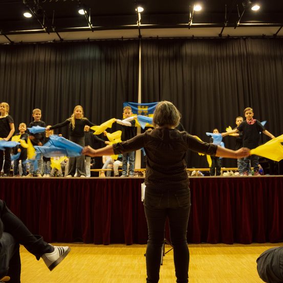 Eine Gruppe von Kindern, die blaue und gelbe Fahnen halten, tritt auf einer Bühne auf, angeführt von einer Frau. Die Bühne ist mit einer roten Tischdecke und einem blauen Hintergrund geschmückt. Scheinwerfer beleuchten die Darsteller.