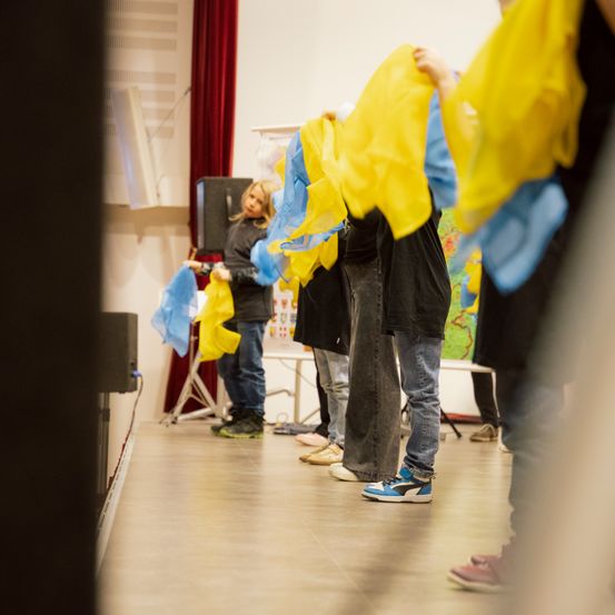 In einem Raum stehen mehrere Personen in einer Reihe und halten gelbe und blaue Banner. Der Raum hat weiße Wände, einen roten Vorhang und einen Lautsprecher. Der Boden ist gefliest.