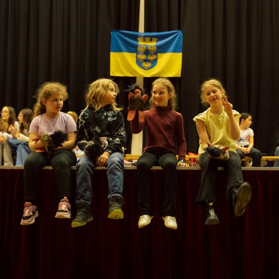 Vier Mädchen sitzen mit Puppen auf der Bühne und lächeln in die Kamera. Hinter ihnen wird eine Flagge gezeigt, und eine Menge sitzt auf Bänken.