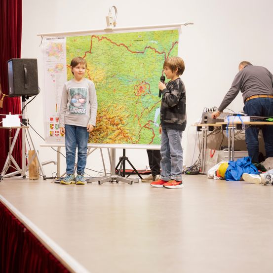 Zwei Jungen auf der Bühne halten Mikrofone. Ein großer Plan ist hinter ihnen. Ein Mann steht neben einem Tisch mit Kabeln.