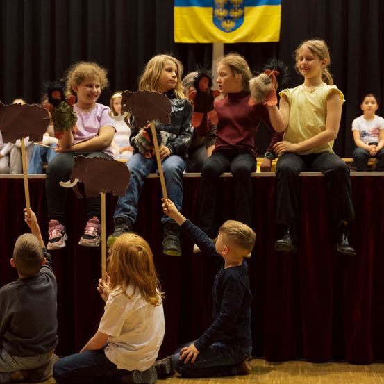 Eine Gruppe von Kindern führt mit Tierpuppen auf der Bühne auf, ein Hintergrund zeigt eine Flagge.