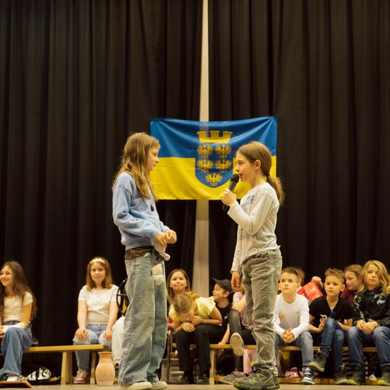 Zwei Mädchen stehen mit einem Mikrofon in der Hand auf der Bühne. Dahinter ist eine ukrainische Flagge zu sehen. Mehrere Kinder sitzen auf Bänken im Publikum.