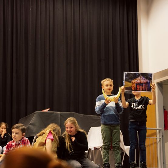 Eine Gruppe von Kindern auf der Bühne, einer hält eine Banane und ein anderer ein Plakat, mit einem schwarzen Vorhang im Hintergrund.