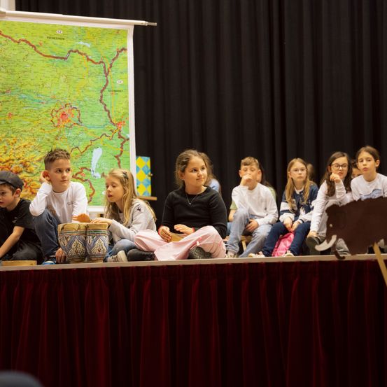 Eine Gruppe von Kindern sitzt auf der Bühne in einem Theater. Sie halten Trommeln und hören aufmerksam zu. Hinter ihnen hängt eine große Karte.